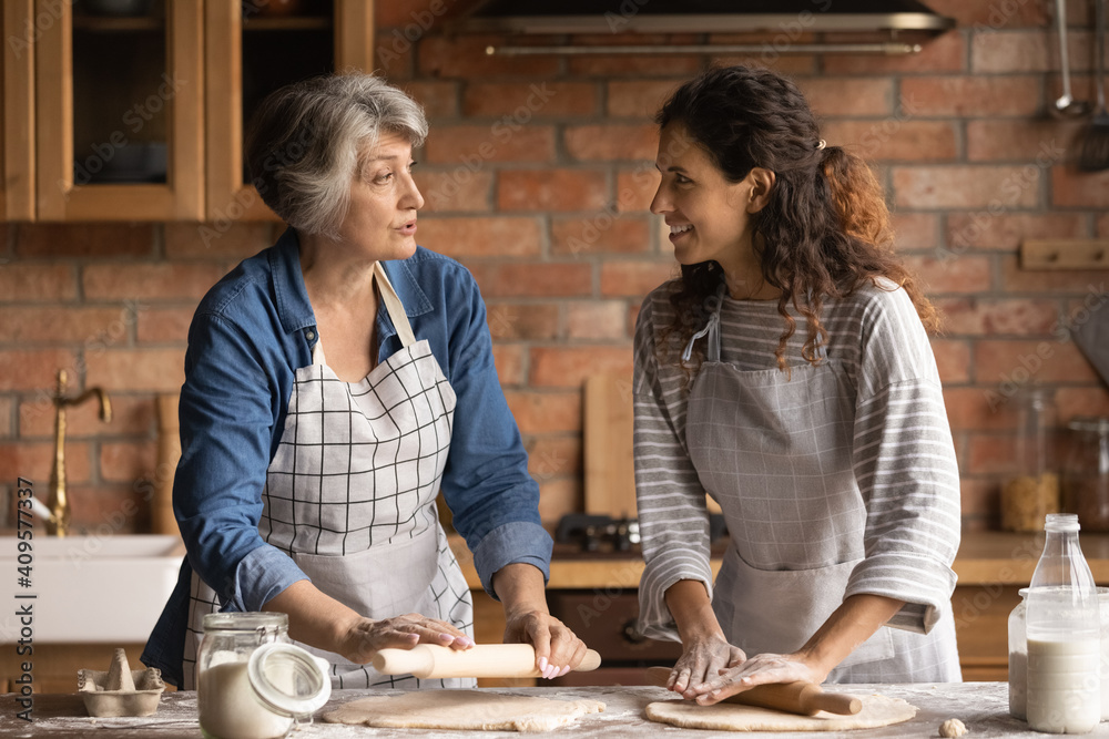 Mature grey haired woman with grownup daughter chatting, cooking homemade pastry, rolling dough, standing in kitchen at home, family spending leisure time together, preparing pie or bread together - obrazy, fototapety, plakaty 
