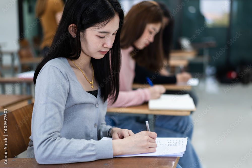 Selective focus of the teen college students sitting on lecture chair in classroom write on examination paper answer sheet in doing the final examination test. Female students in the student uniform. Selective focus of the teen college students sitting on lecture chair in classroom write on examination paper answer sheet in doing the final examination test. Female students in the student uniform.