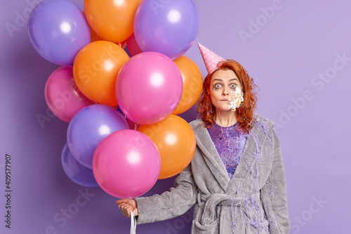Shocked pretty redhead woman has cake on face celebrates graduation from university enjoys free time on party poses with multicolored balloons wears dressing gown and paper cone hat on head.