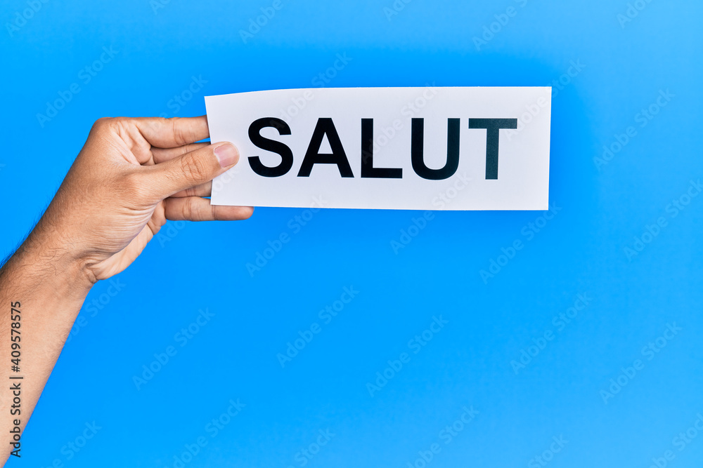 Hand of hispanic man holding salut word paper over isolated blue background. Hand of hispanic man holding salut word paper over isolated blue background.