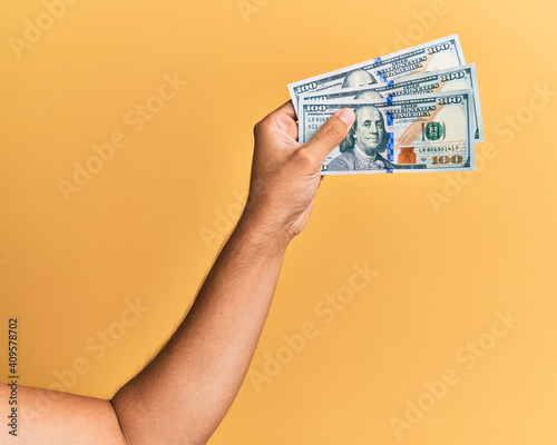 Hand of hispanic man holding 100 dollars banknotes over isolated yellow background.