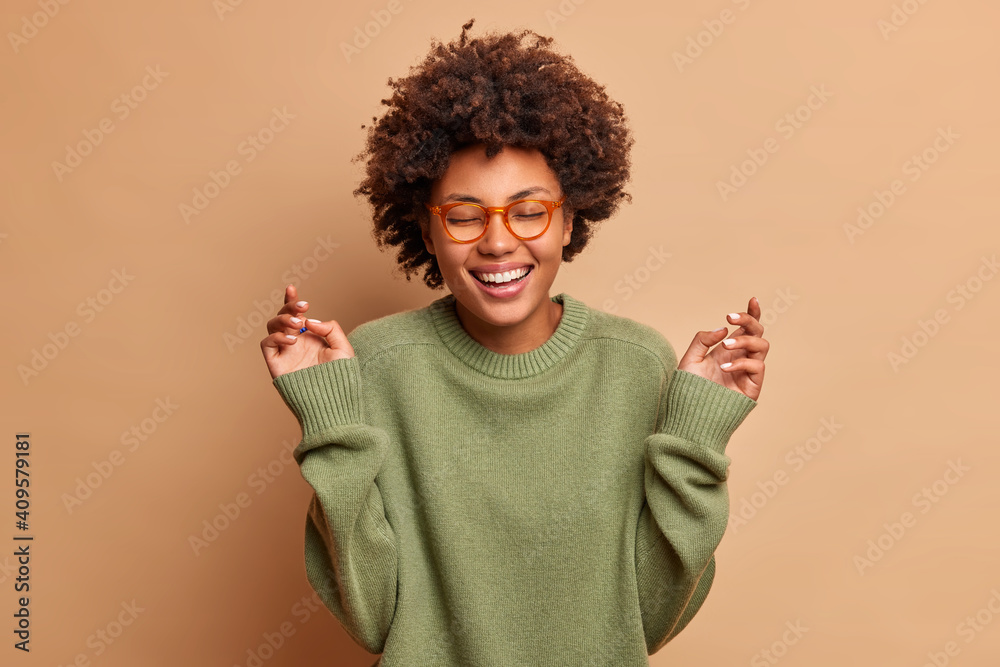 Glad dark skinned woman raises hands has carefree cheerful expression closes eyes smiles toothily wears optical glasses and sweater isolated over beige background. Happiness and joy concept. Glad dark skinned woman raises hands has carefree cheerful expression closes eyes smiles toothily wears optical glasses and sweater isolated over beige background. Happiness and joy concept.
