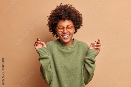 Glad dark skinned woman raises hands has carefree cheerful expression closes eyes smiles toothily wears optical glasses and sweater isolated over beige background. Happiness and joy concept.