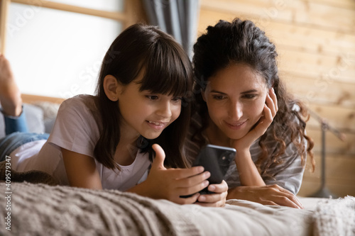 Close up smiling mother and adorable daughter using phone, relaxing on bed at home, curious little girl child with mum looking at smartphone screen, browsing apps, watching video, shopping online