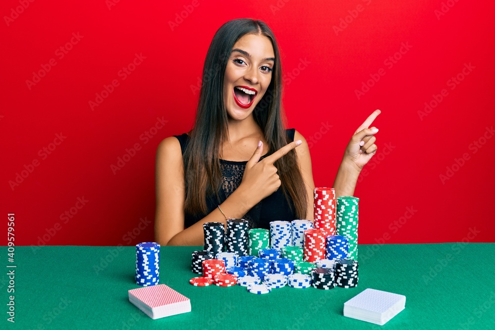 Young hispanic woman sitting on the table playing poker smiling and looking at the camera pointing with two hands and fingers to the side. Young hispanic woman sitting on the table playing poker smiling and looking at the camera pointing with two hands and fingers to the side.