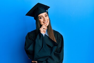 Beautiful brunette young woman wearing graduation cap and ceremony robe looking confident at the camera with smile with crossed arms and hand raised on chin. thinking positive.