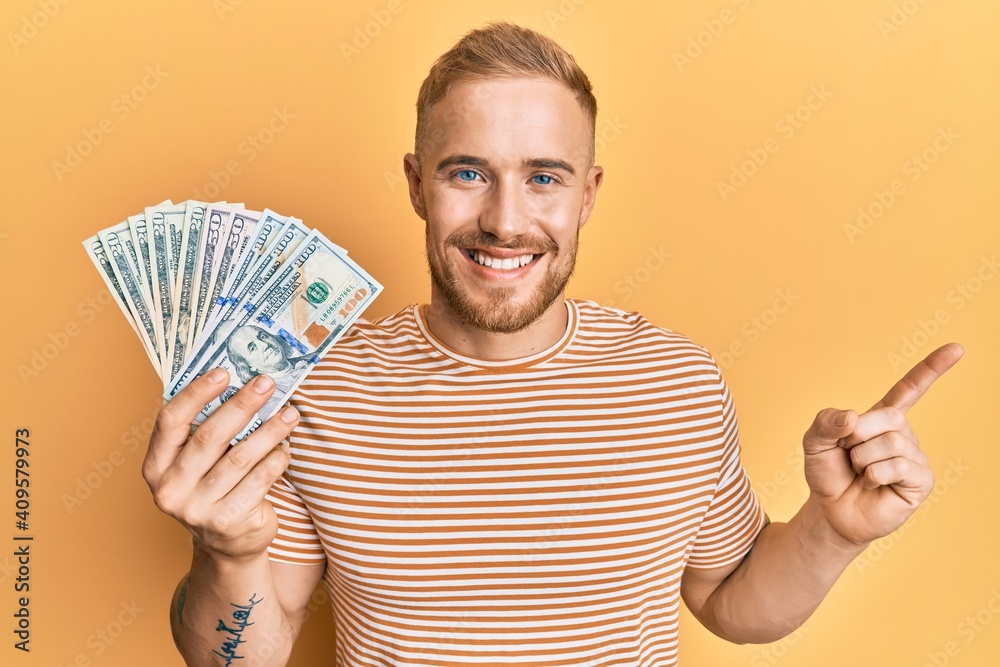 Young caucasian man holding dollars smiling happy pointing with hand and finger to the side Young caucasian man holding dollars smiling happy pointing with hand and finger to the side