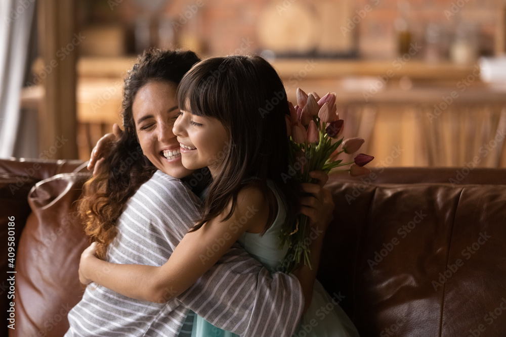 Close up adorable little girl hugging overjoyed young mother, presenting flowers, congratulating with birthday, mothers day or 8 march, family celebrating event, sitting on couch at home together