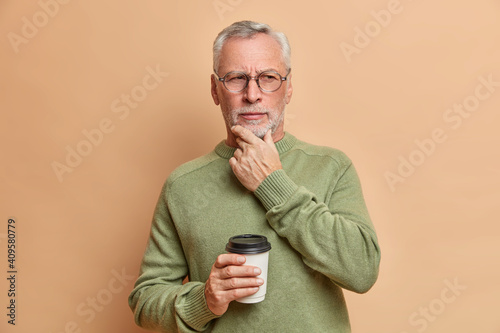 Photo of very serious mature European man holds chin looks pensively aside drinks coffee to go considers something important wears casual sweater and spectacles isolated over brown background © wayhome.studio 