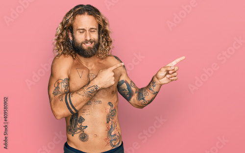 Handsome man with beard and long hair standing shirtless showing tattoos pointing aside worried and nervous with both hands, concerned and surprised expression