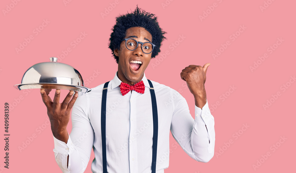 Handsome african american man with afro hair wearing waiter uniform holding silver tray pointing thumb up to the side smiling happy with open mouth Handsome african american man with afro hair wearing waiter uniform holding silver tray pointing thumb up to the side smiling happy with open mouth