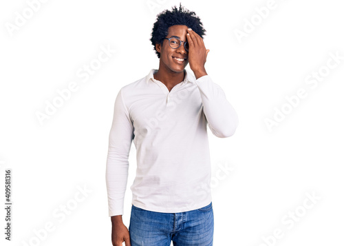 Handsome african american man with afro hair wearing casual clothes and glasses covering one eye with hand, confident smile on face and surprise emotion.