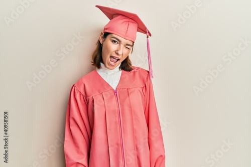 Young caucasian woman wearing graduation cap and ceremony robe winking looking at the camera with sexy expression, cheerful and happy face.