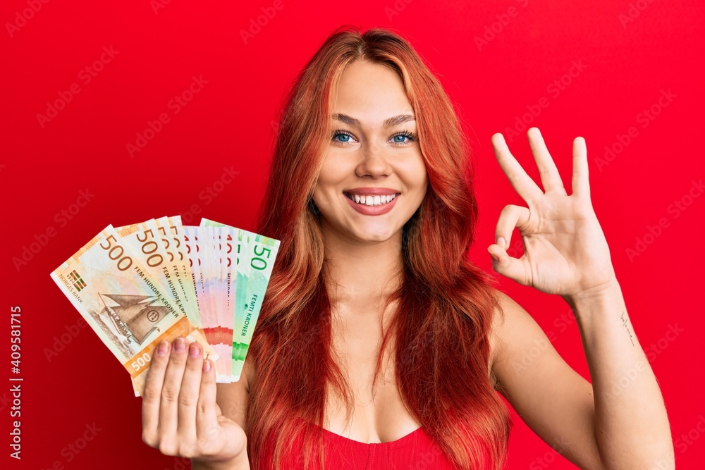 Young beautiful redhead woman holding norwegian krone banknotes doing ok sign with fingers, smiling friendly gesturing excellent symbol Young beautiful redhead woman holding norwegian krone banknotes doing ok sign with fingers, smiling friendly gesturing excellent symbol
