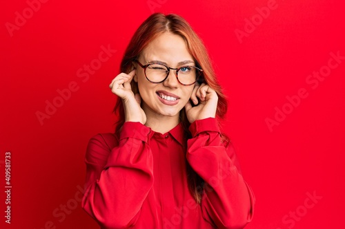 Young beautiful redhead woman wearing casual clothes and glasses over red background covering ears with fingers with annoyed expression for the noise of loud music. deaf concept.
