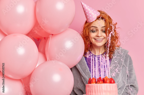 Happy birthday concept. Smiling glad young redhead woman with cheerful makeup and leaked makeup looks gladfully at burning candle on cake makes wish celebrates anniversary holds pink balloons © wayhome.studio 