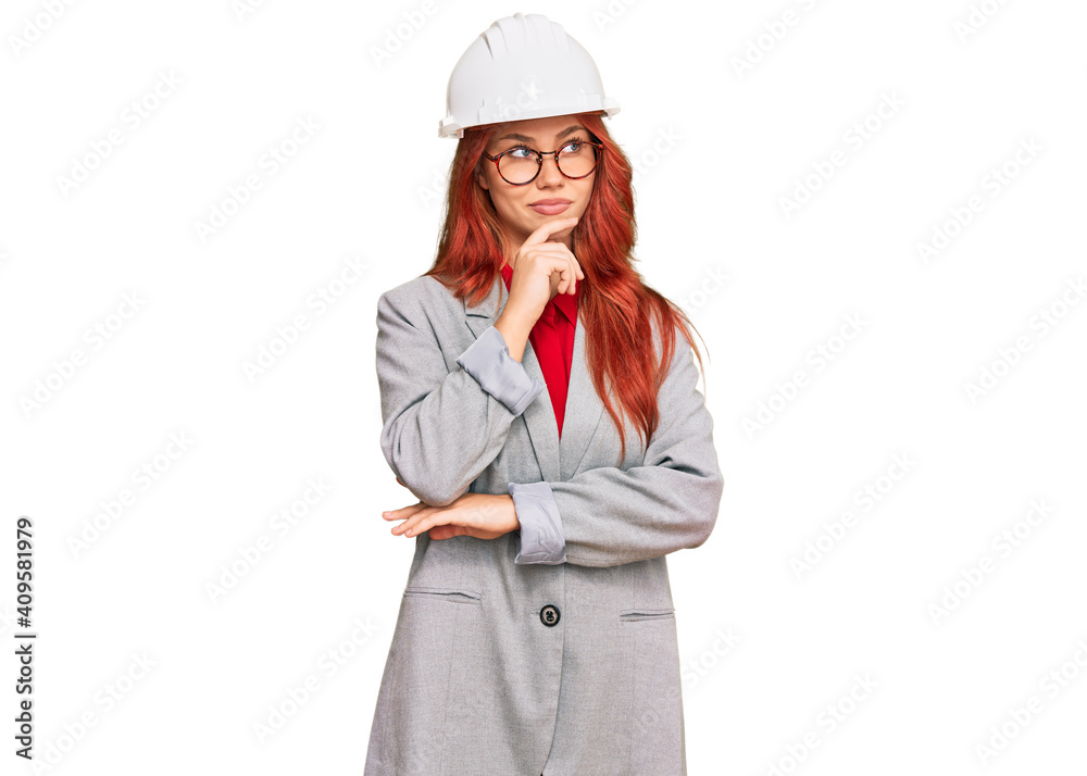 Young redhead woman wearing architect hardhat with hand on chin thinking about question, pensive expression. smiling with thoughtful face. doubt concept.