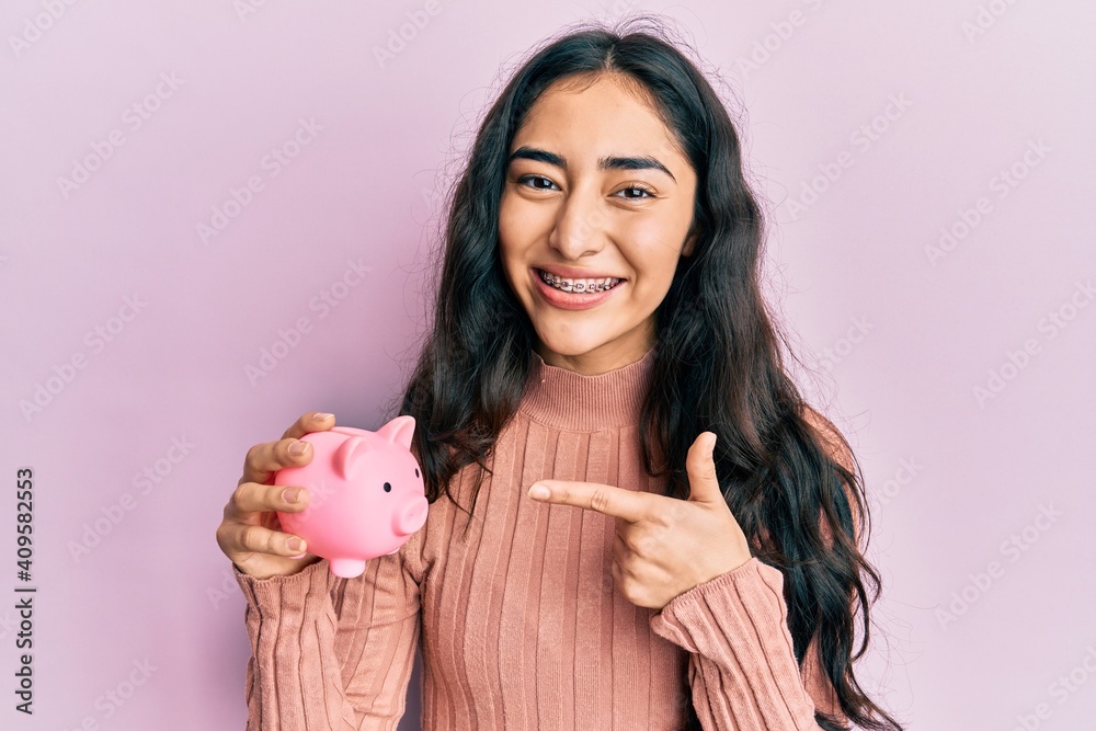 Hispanic teenager girl with dental braces holding piggy bank smiling happy pointing with hand and finger Hispanic teenager girl with dental braces holding piggy bank smiling happy pointing with hand and finger