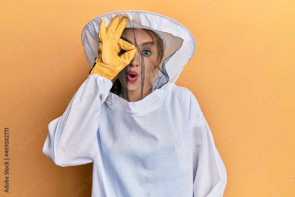Beautiful blonde caucasian woman wearing protective beekeeper uniform doing ok gesture shocked with surprised face, eye looking through fingers. unbelieving expression. Beautiful blonde caucasian woman wearing protective beekeeper uniform doing ok gesture shocked with surprised face, eye looking through fingers. unbelieving expression.