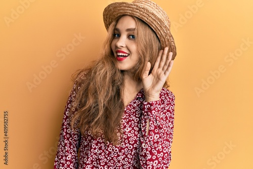 Beautiful blonde caucasian woman wearing summer hat smiling with hand over ear listening an hearing to rumor or gossip. deafness concept.