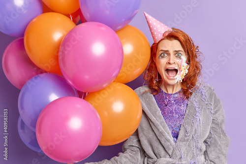 Emotional ginger woman feels annoyed after getting cake in face shouts loudly keeps mouth widely opened celebrates anniversary holds inflated colorful balloons has spoiled makeup. Party time concept