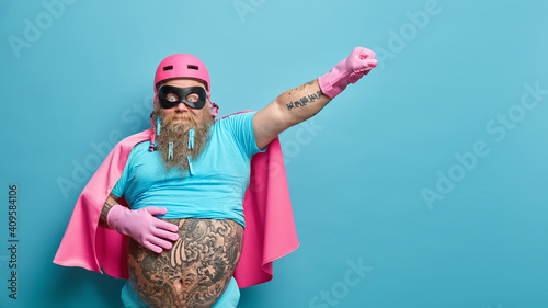 Surprised bearded man dressed like superhero stretches arm as if going to fly keeps hand on big tattooed belly ready for actions isolated over blue background with copy space for your promotion photo