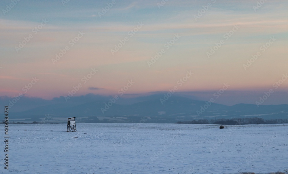 Deerstand on a snowy field during dusk with mountains in the background