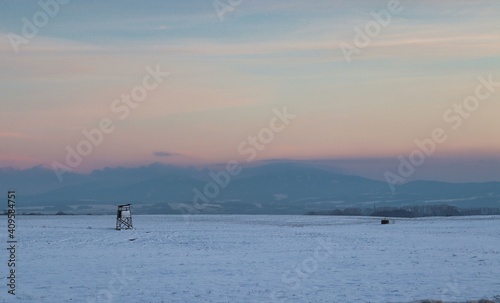 Deerstand on a snowy field during dusk with mountains in the background © CassidyTe