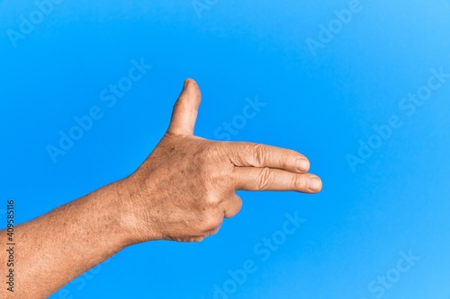 Hand of senior hispanic man over blue isolated background gesturing fire gun weapon with fingers, aiming shoot symbol