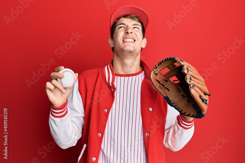 Photography Handsome caucasian man wearing baseball uniform holding golve and ball angry and mad screaming frustrated and furious, shouting with anger looking up