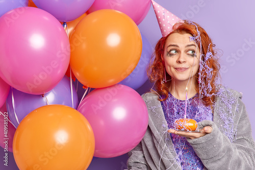 People and holidays concept. Pleased thoughtful redhead young woman looks away wears soft dressing gown and party hat thinks what wish to make before blowing candle poses with colorful balloons