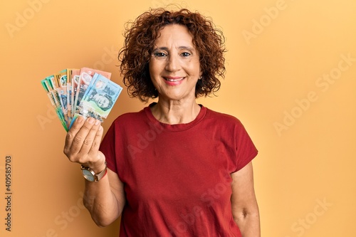 Beautiful middle age mature woman holding australian dollars looking positive and happy standing and smiling with a confident smile showing teeth
