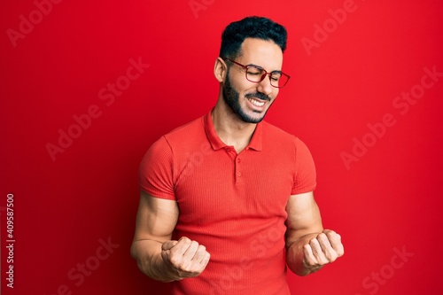 Young hispanic man wearing casual clothes and glasses very happy and excited doing winner gesture with arms raised, smiling and screaming for success. celebration concept.