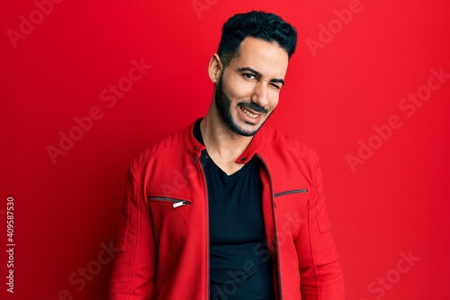 Young hispanic man wearing red leather jacket winking looking at the camera with sexy expression, cheerful and happy face. © Krakenimages.com