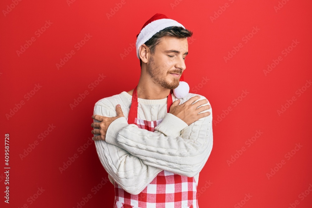 Handsome caucasian man wearing baker apron and christmas hat hugging oneself happy and positive, smiling confident. self love and self care Handsome caucasian man wearing baker apron and christmas hat hugging oneself happy and positive, smiling confident. self love and self care