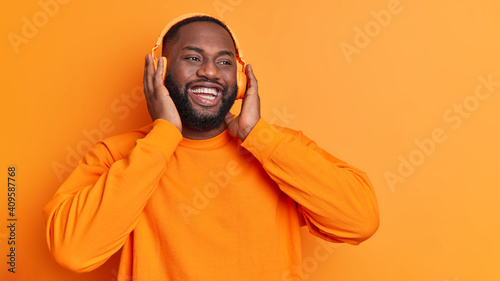 Εκτύπωση καμβά Horizontal shot of happy dark skinned man keeps hands on stereo headphones smile