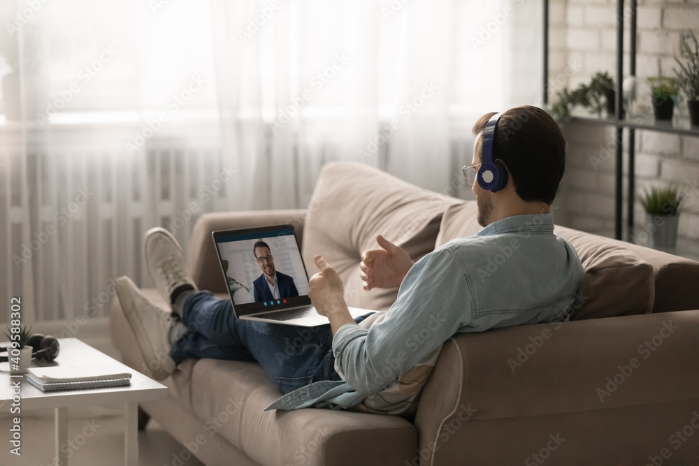 Rear view man wearing headphones using laptop, chatting with colleague online, discussing project strategy, making video call, businessman working at home, sitting on cozy couch in living room