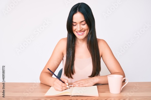 Young caucasian woman sitting at the desk writing book drinking coffee looking positive and happy standing and smiling with a confident smile showing teeth