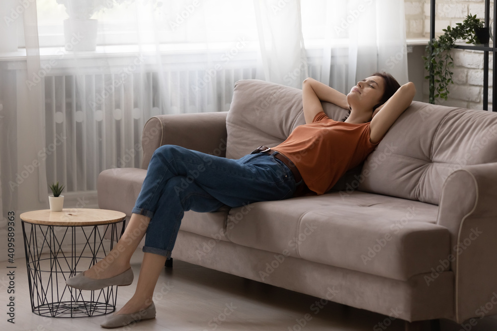 Peaceful young woman relaxing on comfortable couch at home, calm tranquil attractive female taking nap, resting with closed eyes on cozy sofa in modern living room, leaning back, daydreaming