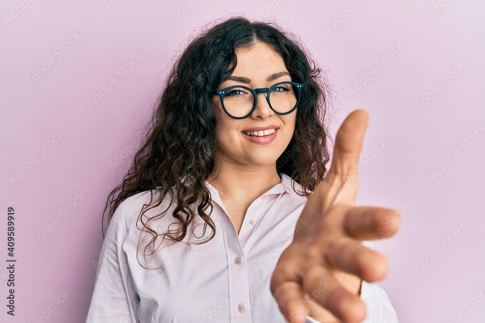 Young brunette woman with curly hair wearing casual clothes and glasses smiling friendly offering handshake as greeting and welcoming. successful business. Young brunette woman with curly hair wearing casual clothes and glasses smiling friendly offering handshake as greeting and welcoming. successful business.