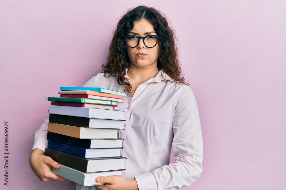 Young brunette woman with curly hair holding a pile of books skeptic and nervous, frowning upset because of problem. negative person. Young brunette woman with curly hair holding a pile of books skeptic and nervous, frowning upset because of problem. negative person.