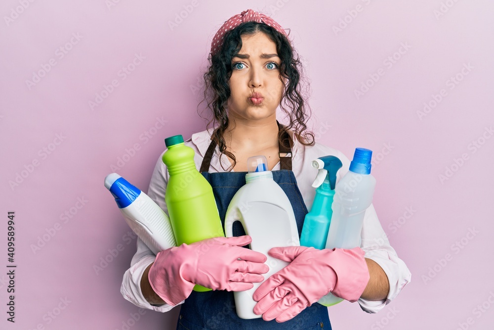Young brunette woman with curly hair wearing cleaner apron holding cleaning products puffing cheeks with funny face. mouth inflated with air, catching air. Young brunette woman with curly hair wearing cleaner apron holding cleaning products puffing cheeks with funny face. mouth inflated with air, catching air.