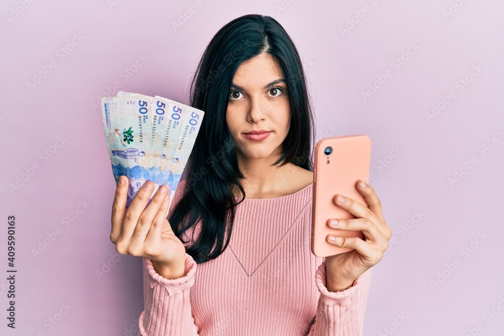 Young hispanic woman using smartphone holding colombian pesos banknotes relaxed with serious expression on face. simple and natural looking at the camera.