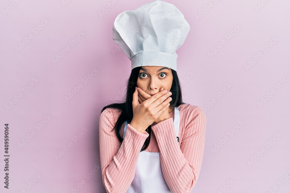 Young hispanic woman wearing baker uniform and cook hat shocked covering mouth with hands for mistake. secret concept. Young hispanic woman wearing baker uniform and cook hat shocked covering mouth with hands for mistake. secret concept.