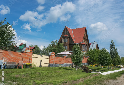 A street in the village of Sedelnikovo (Ural, Russia) with a beautiful tall wooden house in the form of a tower, a fence and a green lawn with flower beds and a bench. Lifestyle, care and relaxation 