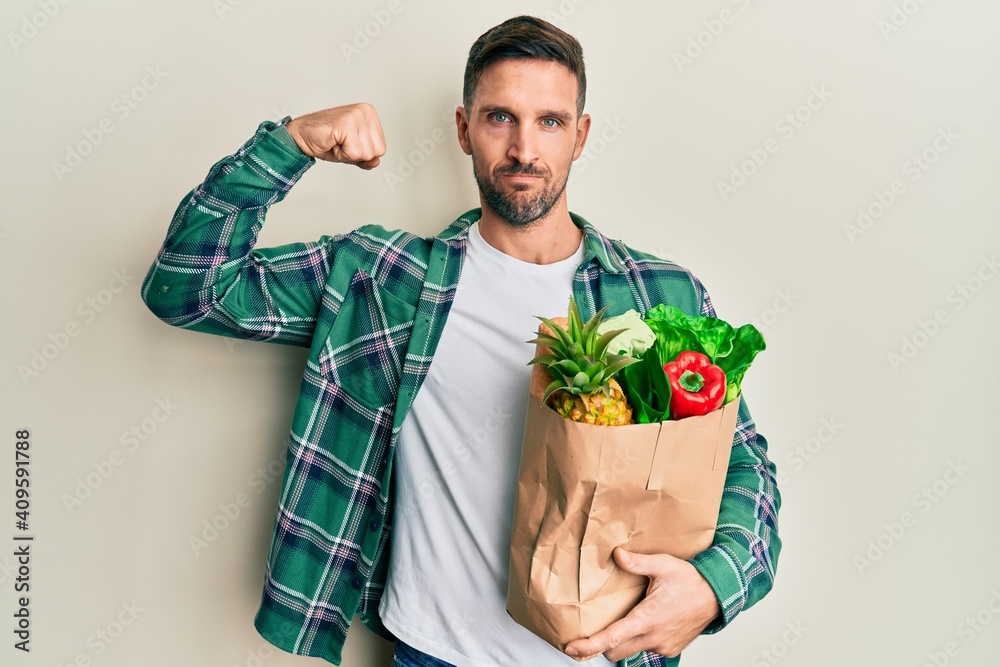 Handsome man with beard holding paper bag with groceries strong person showing arm muscle, confident and proud of power - obrazy, fototapety, plakaty 