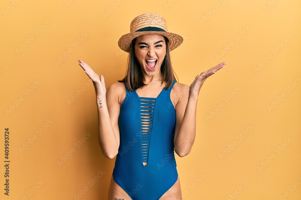 Young brunette girl wearing swimsuit and summer hat celebrating mad and crazy for success with arms raised and closed eyes screaming excited. winner concept Young brunette girl wearing swimsuit and summer hat celebrating mad and crazy for success with arms raised and closed eyes screaming excited. winner concept