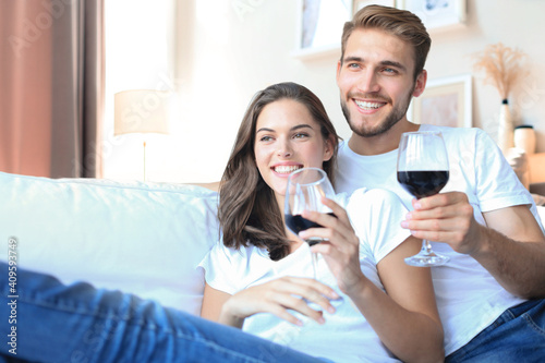 Happy couple sitting, relaxing on couch in living room, using laptop for online shopping together, watching movie, drinking red wine.