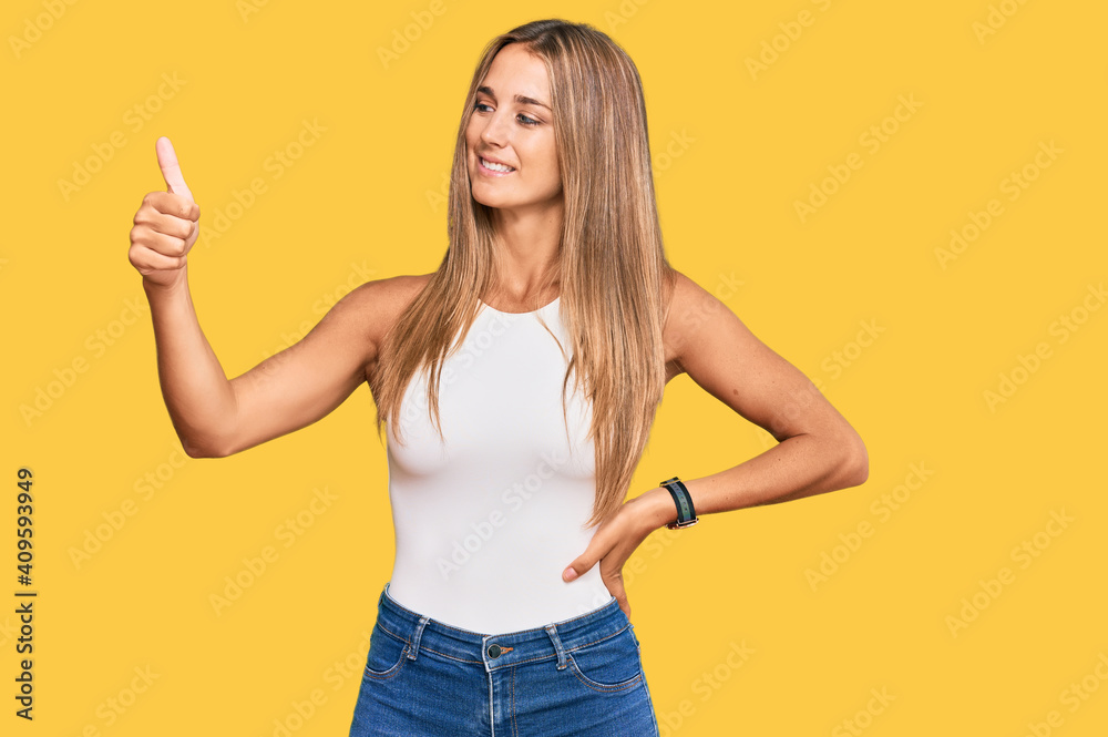 Young blonde woman wearing casual style with sleeveless shirt looking proud, smiling doing thumbs up gesture to the side Young blonde woman wearing casual style with sleeveless shirt looking proud, smiling doing thumbs up gesture to the side
