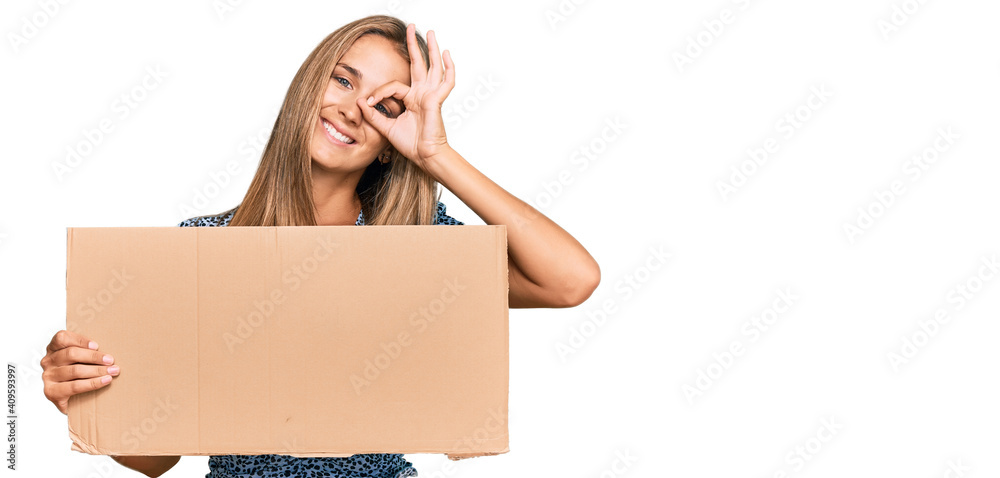 Young blonde woman holding cardboard empty banner smiling happy doing ok sign with hand on eye looking through fingers Young blonde woman holding cardboard empty banner smiling happy doing ok sign with hand on eye looking through fingers